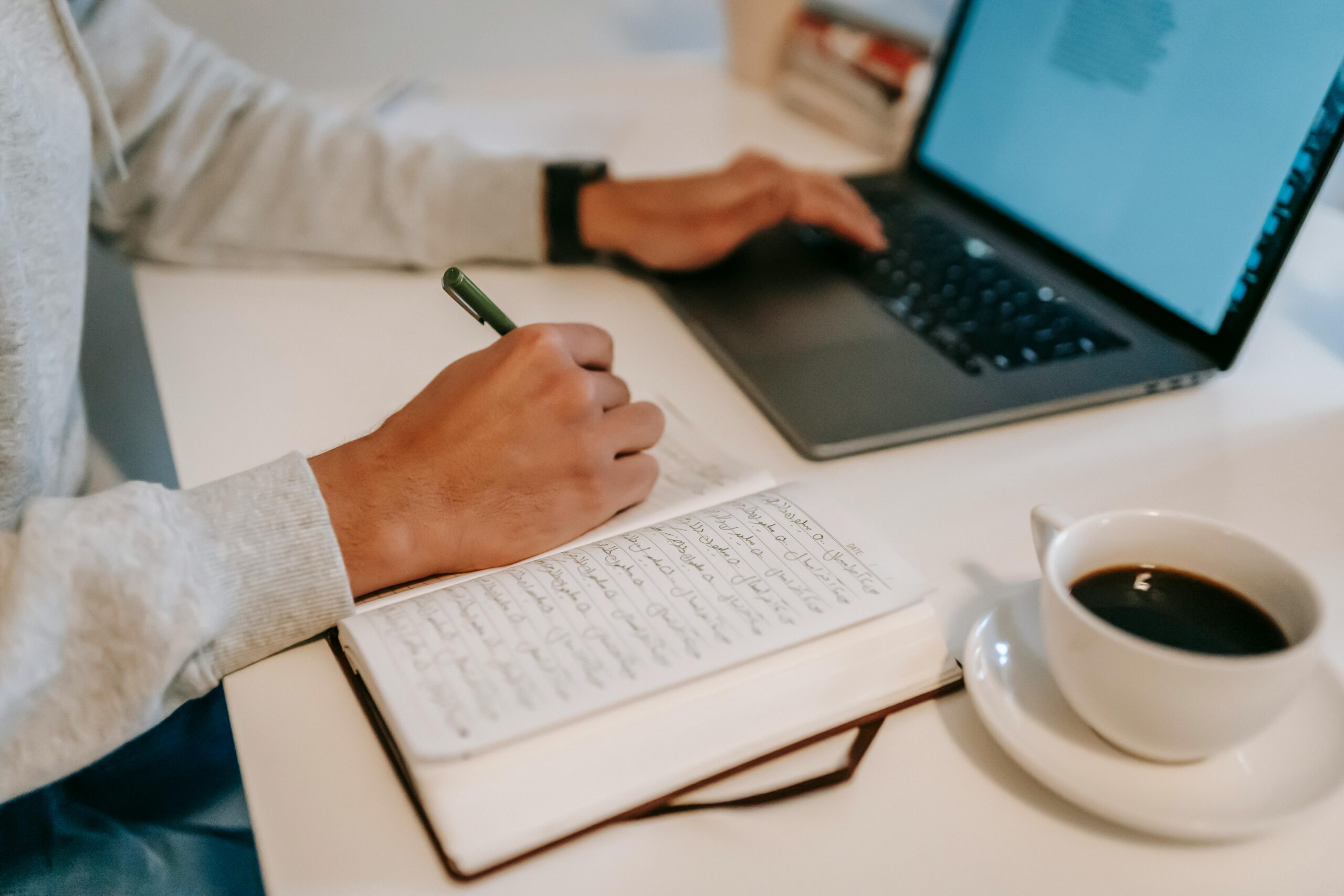 A close-up of a person writing in a notebook while using a laptop, accompanied by a cup of coffee.